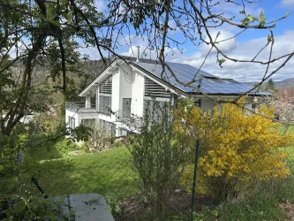 Ein modernes Haus mit Solarpaneelen steht umgeben von grünem Gras und bunten Sträuchern. Im Hintergrund sind einige Wolken am Himmel sichtbar.