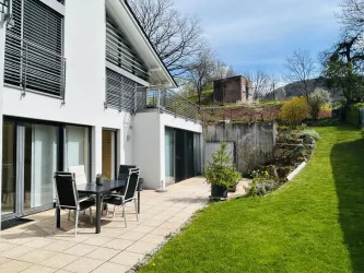 Eine moderne Terrasse mit einem Esstisch und Stühlen. Im Hintergrund befindet sich ein gepflegter Garten und ein grüner Rasen.