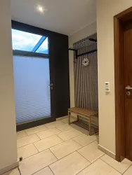 Ein moderner Eingangsbereich mit hellen Fliesen und einer Holzbank. Ein Fenster sorgt für Tageslicht und macht den Raum einladend.