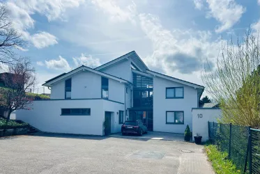 Ein modernes Haus mit einem klaren Design und großen Fenstern. Der Parkplatz ist vor dem Gebäude sichtbar, umgeben von grüner Vegetation.