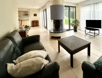 Ein modernes Wohnzimmer mit einem großen Fenster und Blick auf den Garten. Es gibt ein komfortables Sofa, einen Tisch und einen Fernseher.