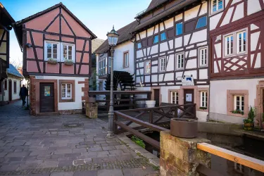 Ein malerischer Platz mit Fachwerkhäusern in hübschen Farben. Im Hintergrund ist ein kleiner Wasserlauf und eine Straße zu sehen.