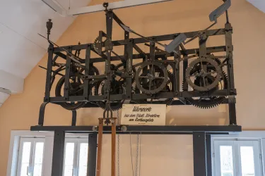 Eine historische mechanische Uhr mit sichtbaren Zahnrädern und einem Rahmen. Die Uhr hängt an der Wand in einem hellen Raum.