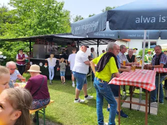 Eine lebendige Gartenparty mit vielen Menschen, die an Tischen sitzen und trinken. Es gibt einen Foodstand im Hintergrund und viel Grün um die Veranstaltung herum.