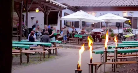 Ein Außenbereich mit Holzbankreihen und Tischen. Gäste genießen die Atmosphäre unter Sonnenschirmen, während Fackeln flammen.