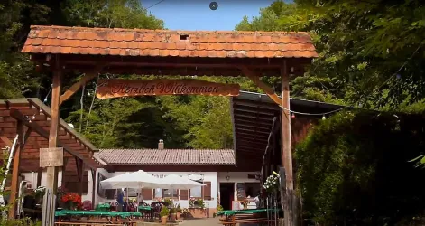 Ein einladendes Holztor mit der Aufschrift "Herzlich Willkommen". Im Hintergrund ist ein Gasthaus umgeben von Bäumen und Sitzplätzen im Freien zu sehen.