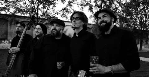 Eine Musikgruppe steht im Freien unter Bäumen. Alle Mitglieder tragen schwarze Kleidung und lächeln in die Kamera.