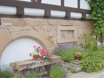 Ein stiller Außenbereich mit einer steinernen Bank und blühenden Pflanzen. Die Wand ist aus hellem Stein mit einem traditionellen Holzrahmen.