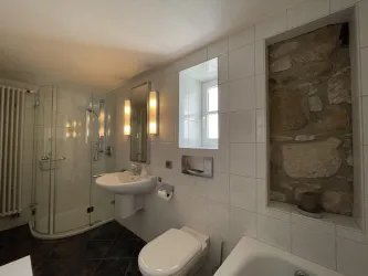 Ein modernes Badezimmer mit weißer Fliesenwand und einem großen Fenster. Es enthält einen Duschbereich, ein Waschbecken und eine Badewanne.