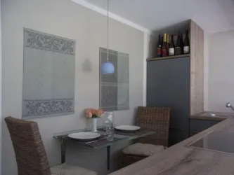 Eine modern eingerichtete Küche mit einem kleinen Essbereich. Auf dem Tisch stehen Geschirr und eine Blumenvase, und im Hintergrund sind Flaschen in einem Regal sichtbar.