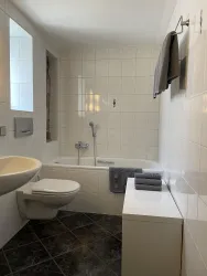 Ein modernes Badezimmer mit einer Badewanne und einer Toilette. Die Wände sind hell gefliest und es gibt Handtücher und Toilettenartikel.