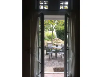 Eine offene Tür mit Blick auf einen schönen Garten. Draußen stehen ein Tisch und Stühle auf einem gepflasterten Platz.