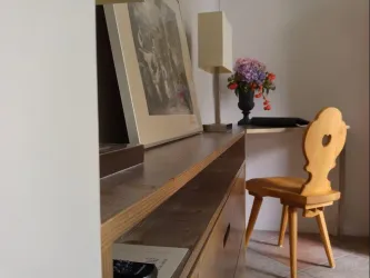 Ein stilvolles Zimmer mit einem Holzmöbel, einem Bild und einer Lampe. Auf dem Tisch steht eine Vase mit bunten Blumen und ein Holzstuhl mit einer herzförmigen Rückenlehne.