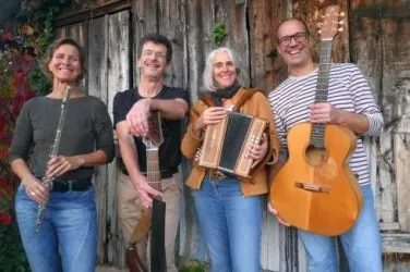 Eine Gruppe von vier Musikern steht lächelnd vor einer alten Holzwand. Sie halten verschiedene Musikinstrumente, darunter eine Flöte, eine Gitarre und ein Akkordeon.