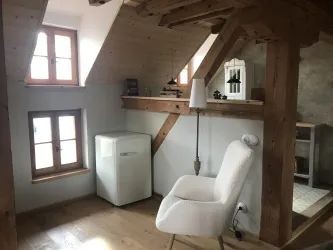 Ein gemütliches Zimmer mit Holzdecken und großen Fenstern. Es enthält einen Sessel, einen Kühlschrank und dekorative Elemente.