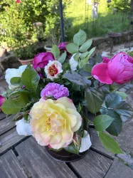Ein bunter Blumenstrauß mit rosa und gelben Blumen steht auf einem Holztisch. Im Hintergrund sind grüne Pflanzen zu sehen.