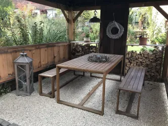 Ein gemütlicher Außenbereich mit einem Holztisch und zwei Bänken. Im Hintergrund sind Pflanzen und eine Laterne zu sehen.