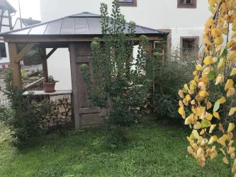 Ein Garten mit einem Holzpavillon und grünem Gras. Im Vordergrund sind orangefarbene Blätter eines Baumes zu sehen.