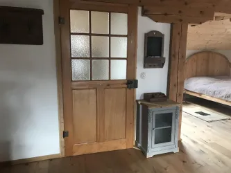 Ein gemütlicher Raum mit einer Holztür und einem Fenster. Neben der Tür steht ein kleiner Ofen und im Hintergrund ist ein Bett sichtbar.