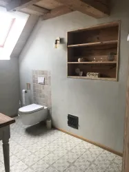 Ein modernes Badezimmer mit schlichten Wänden und einem schrägen Dach. Es enthält eine Toilette, ein Regal aus Holz und einen schönen Fliesenboden.