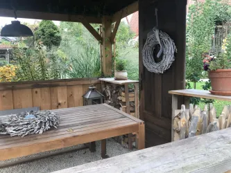 Eine gemütliche Holzterrasse mit einem Tisch und einer Holzdeko. Im Hintergrund sind Pflanzen und eine dekorative Laterne zu sehen.