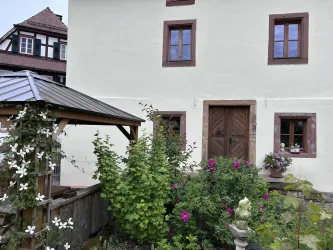 Ein charmantes Haus mit einem schlichten weißen Anstrich und braunen Fensterrahmen. Im Vordergrund sind blühende Pflanzen und ein Pavillon zu sehen.