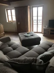 Ein gemütlicher Wohnbereich mit einem großen, grauen Sofa und einem Tisch in der Mitte. Die Fenster lassen viel Tageslicht herein und bieten einen Blick nach draußen.