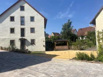Ein großes, helles Haus steht in einer ruhigen Umgebung. Im Vordergrund ist ein gepflasterter Weg zu sehen, neben einem Garten mit einer Pergola und viel Grün.