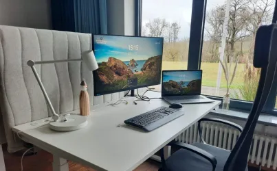 Ein moderner Schreibtisch mit zwei Bildschirmen, einer Lampe und einer Tasse. Im Hintergrund sind große Fenster mit Blick auf eine grüne Landschaft.