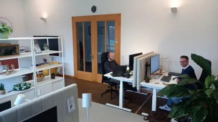 Ein modernes Büro mit zwei Schreibtischen und Computern. Im Hintergrund sind Regale mit Pflanzen und Büromaterialien zu sehen.