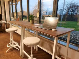 Ein moderner Arbeitsplatz mit einem langen Tisch und hellen Stühlen. Ein Laptop steht auf dem Tisch, während große Fenster den Blick ins Grüne freigeben.