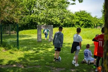 Ein Gruppen von Kindern spielt auf einer Wiese. Im Hintergrund steht ein Tor aus Steinen und Bäume umgeben den Platz.