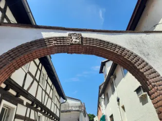 Ein alter, bogenförmiger Eingang aus Ziegelsteinen, der zwischen zwei Häusern hindurchführt. Darüber ist ein Wappen in das Mauerwerk eingearbeitet, und der Himmel ist klar und blau.