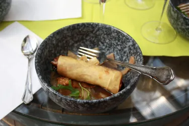 Ein tiefes, graues Schalen mit einem Frühlingsroll in Brühe und frischen Kräutern. Gabel und Löffel liegen neben dem Teller.