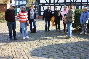 Eine Gruppe von Menschen steht auf einem gepflasterten Platz. Im Hintergrund sind Fachwerkhäuser und frisches Grün zu sehen.
