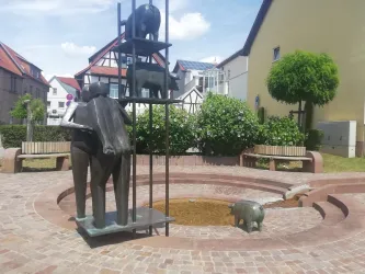 Eine moderne Skulptur mit Figuren und Tieren in einem Park. Rund um die Skulptur stehen Sitzbänke und grüne Sträucher.