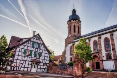 Eine historische Stadtansicht mit einem Fachwerkhaus und einer Kirche mit hohem Turm. Der Himmel ist klar und zeigt einige Kondensstreifen.