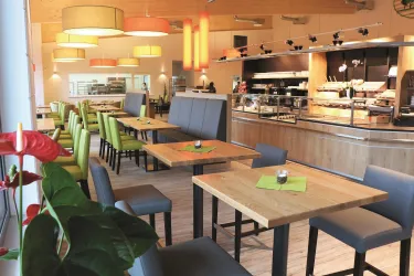 Ein modernes Restaurant mit Holzdekor und bunten Lampen. Die Tische sind mit grünen und grauen Stühlen gedeckt und laden zum Verweilen ein.