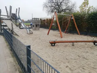 Ein Spielplatz mit verschiedenen Spielgeräten, darunter eine Rutsche und eine Schaukel. Der Boden ist mit Sand bedeckt und von einem Zaun umgeben.