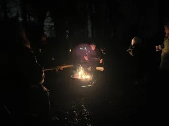 Eine Gruppe von Menschen sitzt im Dunkeln um ein Lagerfeuer. Die Atmosphäre ist gemütlich und einladend.