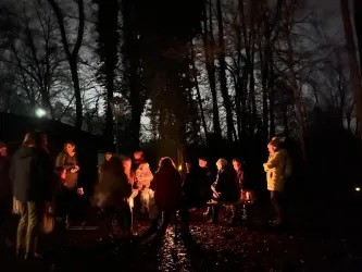 Eine Gruppe von Menschen sitzt um ein Lagerfeuer in der Dämmerung. Die Umgebung ist von dunklen Bäumen umgeben und der Himmel ist leicht bewölkt.