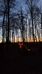 Zwei Personen stehen im Wald bei Dämmerung und feuern ein Lagerfeuer an. Der Himmel ist in warmen Farben während die Sonne untergeht.