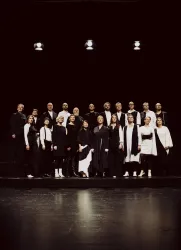 Eine Gruppe von Sängern steht auf einer Bühne, alle in schwarz und weiß gekleidet. Der Hintergrund ist dunkel, und es gibt ein paar Scheinwerfer, die die Gruppe beleuchten.
