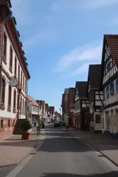 Eine ruhige Straße mit historischen Fachwerkhäusern und klarem Himmel. Es sind einige Menschen und Autos sichtbar.