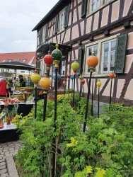 Eine malerische Szene mit einem Fachwerkhaus im Hintergrund. Bunte Zapfen stehen in einem Garten mit grünen Pflanzen.
