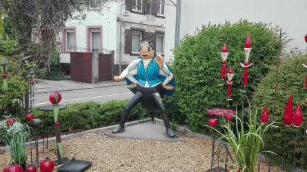 Eine auffällige Figur eines kräftigen Mannes im Garten steht zwischen bunten Pflanzen und Dekorationen. Im Hintergrund sind Gebäude und eine Straße sichtbar.