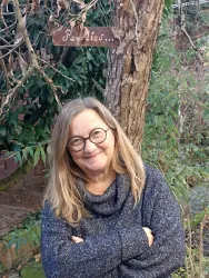 Eine lächelnde Frau steht vor einem Baum mit einer Holztafel im Hintergrund. Die Umgebung ist grün und lebendig.