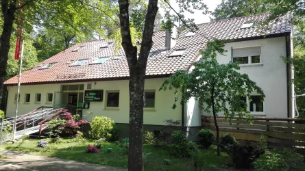 Ein modernes Gebäude umgeben von Bäumen und Blumen. Das Haus hat einen roten Dacht und große Fenster.