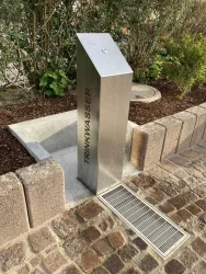 Ein Edelstahl-Wasserbrunnen steht im Freien. Er ist mit dem Wort "TRINKWASSER" beschriftet und umgeben von Pflanzen und einem Pflastersteinweg.