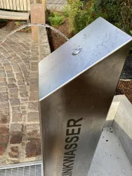 Ein moderner Trinkbrunnen aus Edelstahl mit fließendem Wasser. Er steht in einer grünen Umgebung neben einem gepflasterten Weg.